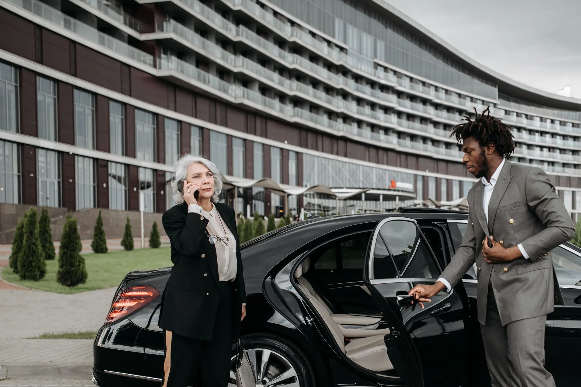 A chauffeur in a dark suit opens the rear door of a black luxury car.